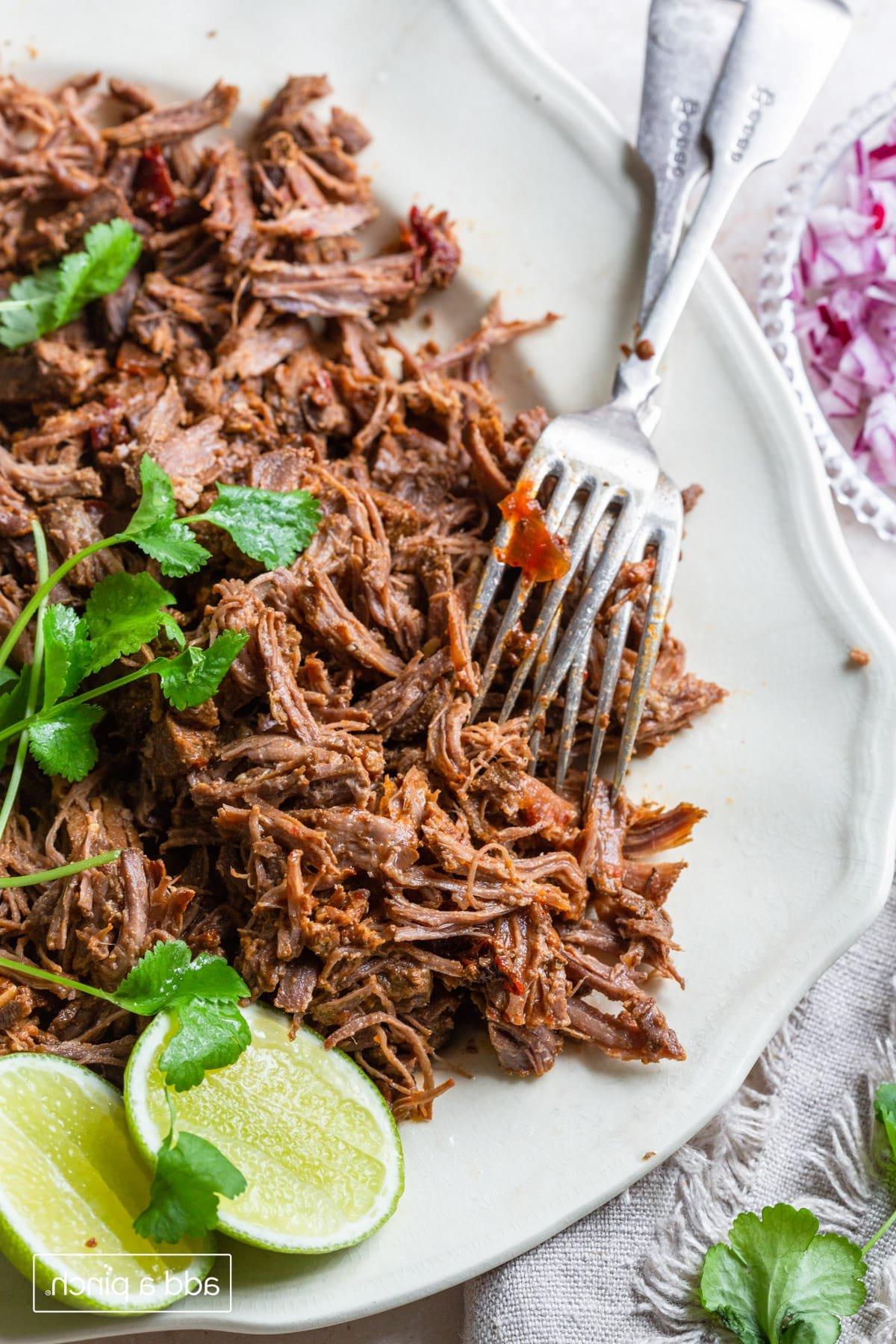 Εύκολη Συνταγή Μπαρμπακόα 8 Barbacoa σε πιατέλα με κόλιανδρο, φέτες λάιμ και δύο πιρούνια.
