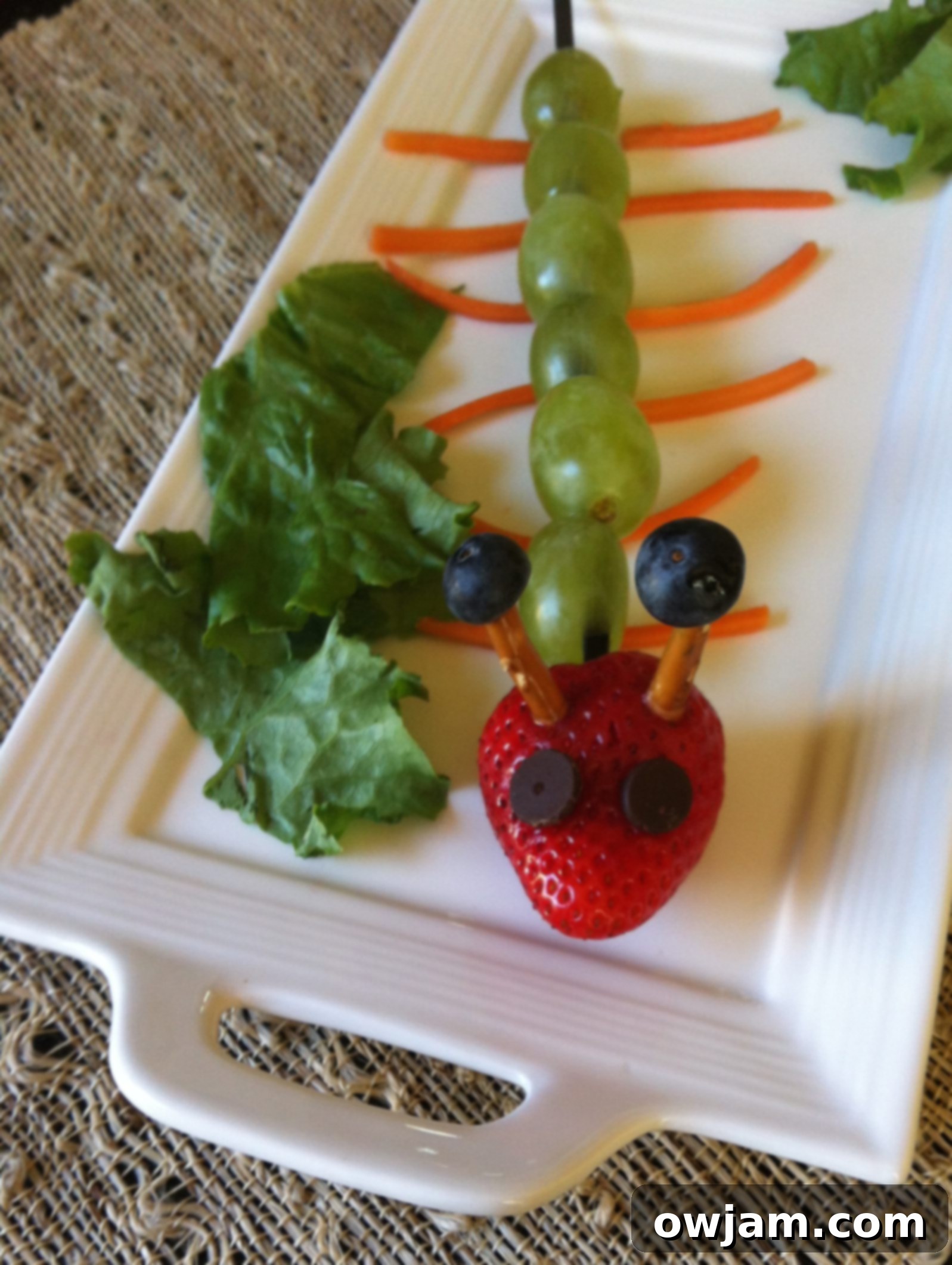 A close-up of the fruit caterpillar on a white plate, highlighting the grape body and strawberry head with chocolate chip eyes.
