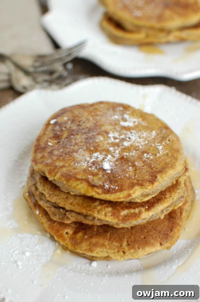 A beautifully stacked plate of fluffy Pumpkin Pie Pancakes, generously drizzled with maple syrup and powdered sugar, ready for a delicious fall breakfast.