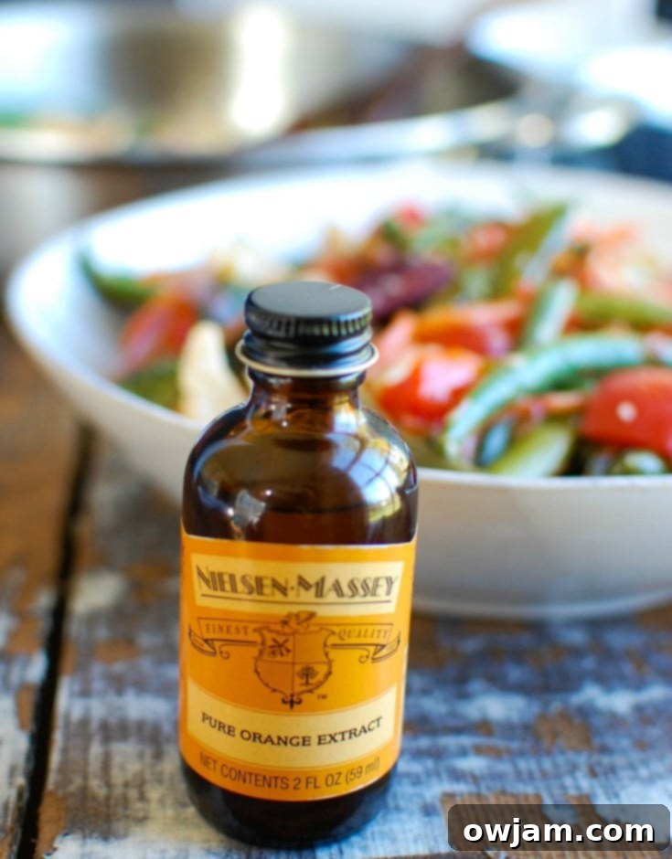 A flat lay shot showcasing the fresh ingredients for Orange Chicken Stir Fry, including a bottle of Nielsen-Massey Pure Orange Extract, chicken breast, various chopped vegetables, and seasoning elements.