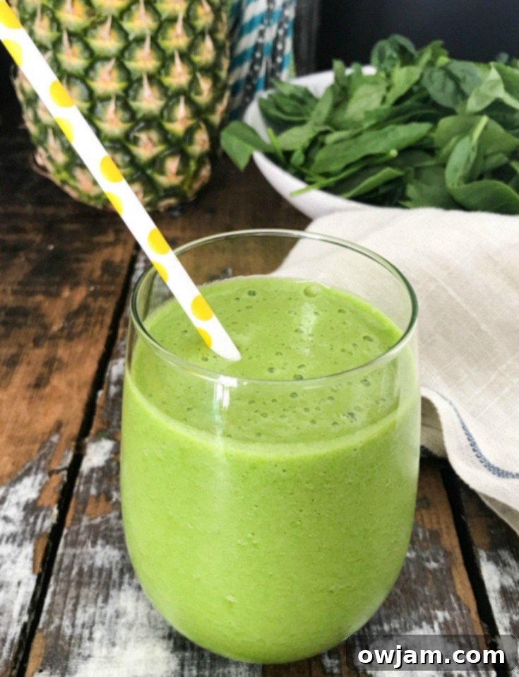 A glass of Tropical Green Smoothie next to a pile of fresh spinach, pineapple chunks, Granny Smith apple, and mint leaves, highlighting the main ingredients.