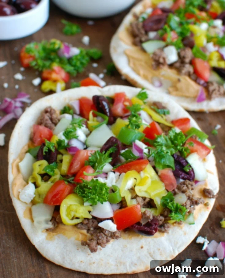 Fresh ingredients for Greek Beef Pita Pizza laid out on a cutting board, including red onion, tomatoes, cucumbers, and feta.