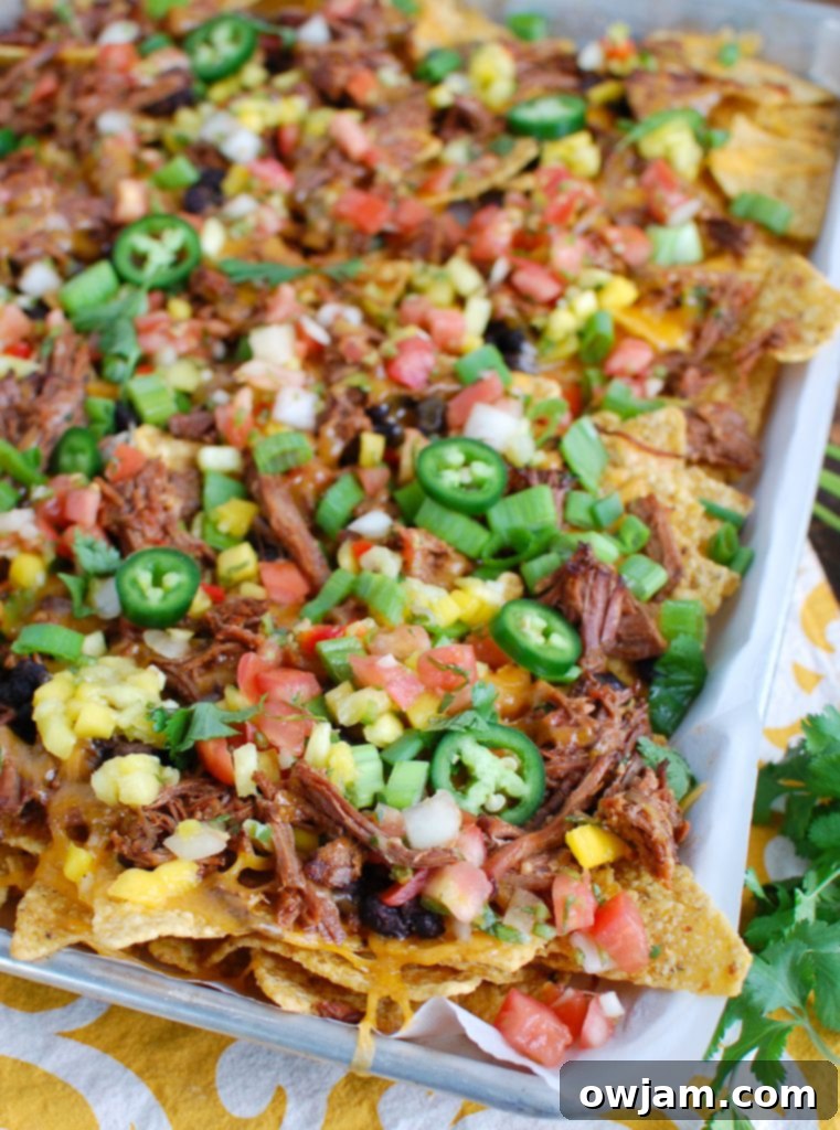 Sheet pan loaded with homemade beef nachos, topped with slow cooker shredded beef, black beans, salsa, jalapeños, cheese, green onions, and cilantro.