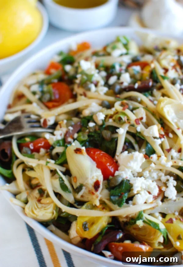 Sun-Kissed Mediterranean Pasta 2 White bowl of Mediterranean Pasta with a fork, showcasing fresh ingredients like halved cherry tomatoes, olives, crumbled feta cheese, and green spinach.