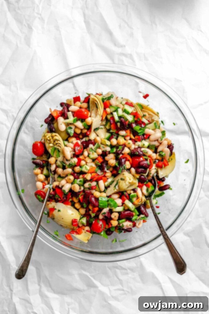 Close-up of a bowl filled with colorful Mediterranean Three Bean Salad ingredients, being tossed with two spoons.