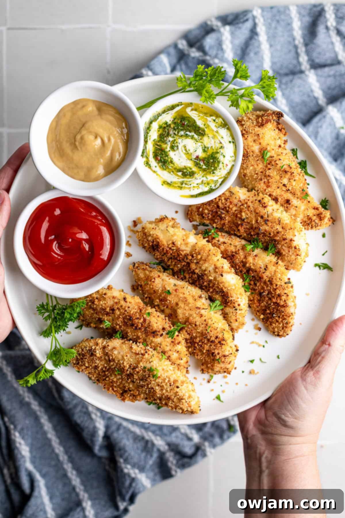 Super Crispy Easy Baked Chicken Tenders 2 Crispy baked chicken fingers on a white plate, golden brown and ready to serve with three dipping sauces, garnished with fresh parsley.