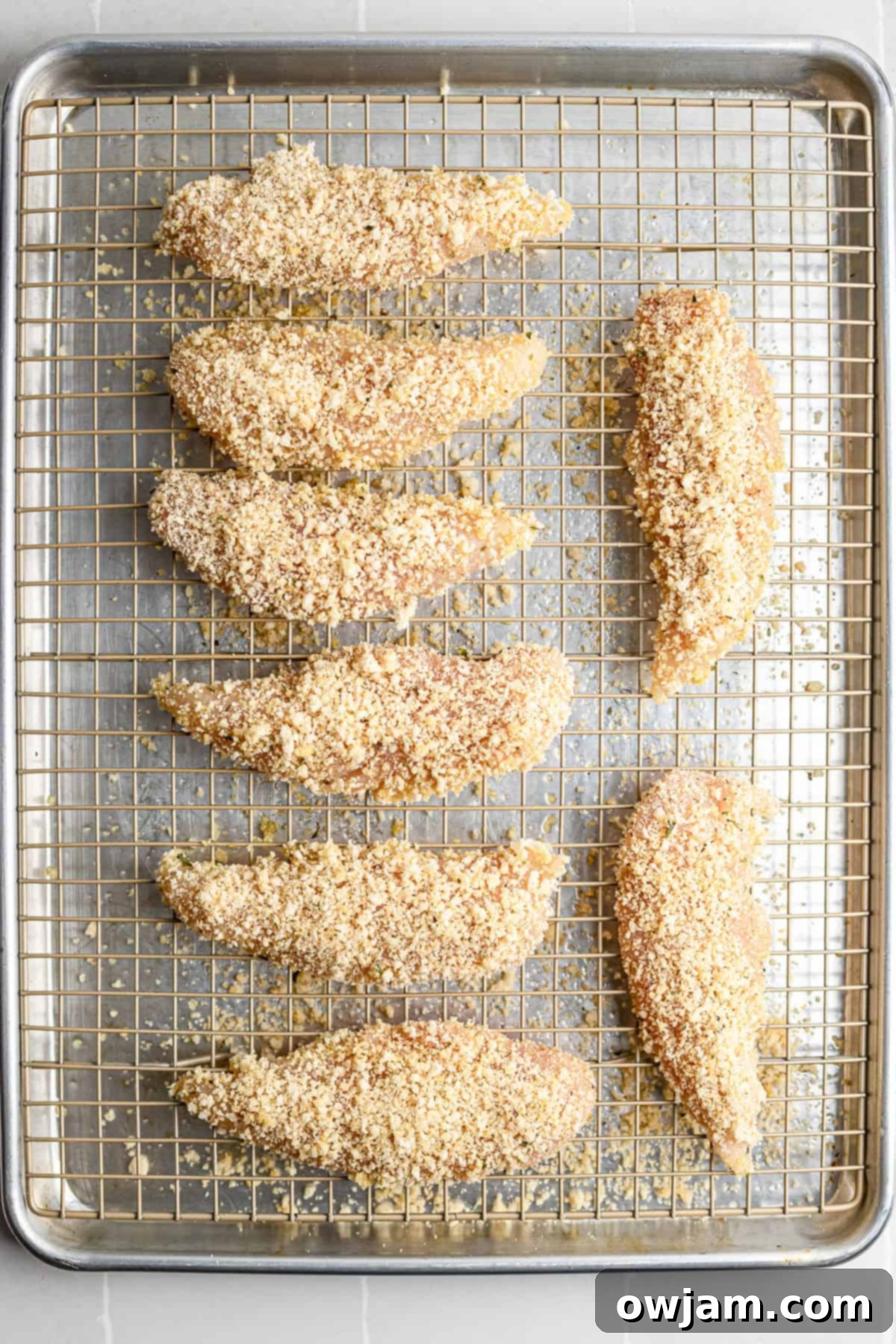 Super Crispy Easy Baked Chicken Tenders 6 Breaded chicken tenders arranged neatly on a wire rack over a baking sheet, ready for the oven.