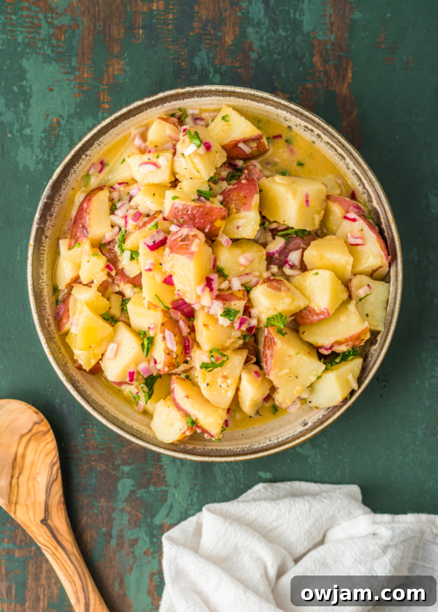 Warm German Potato Salad in a serving bowl with a wooden spoon, garnished with fresh herbs.