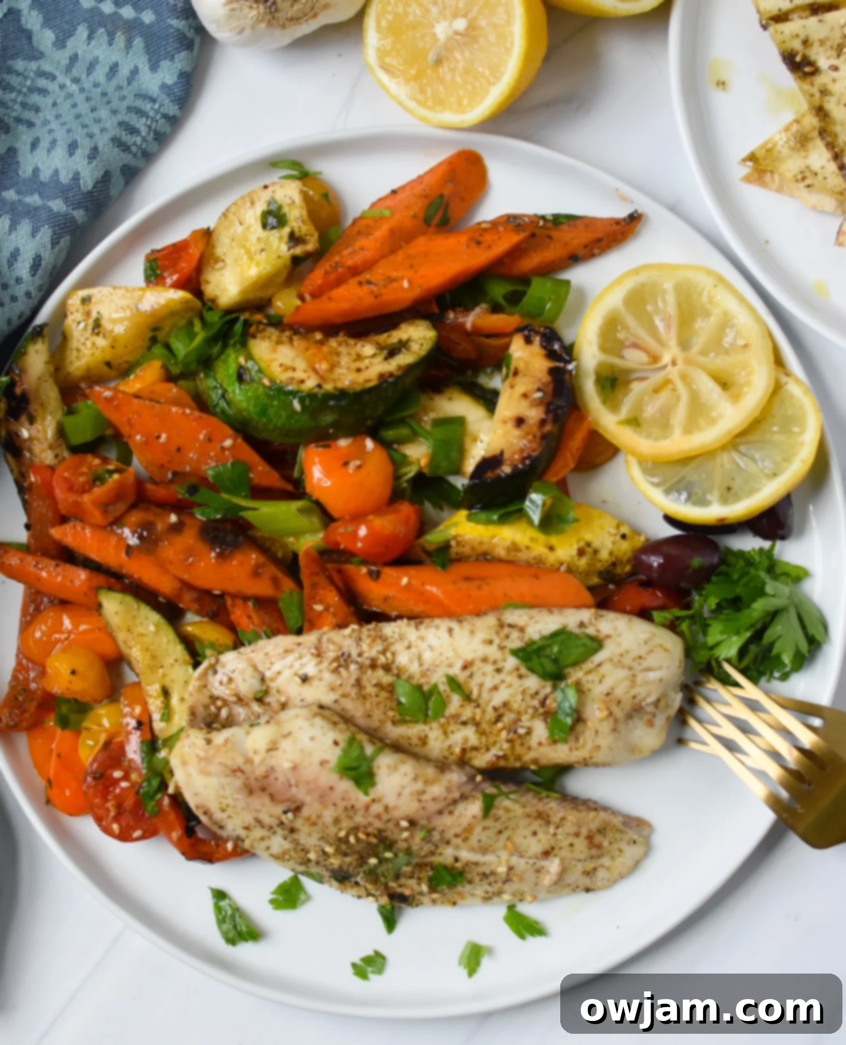 A single serving of Mediterranean Baked Tilapia with roasted vegetables and a lemon wedge on a plate, garnished with fresh herbs.