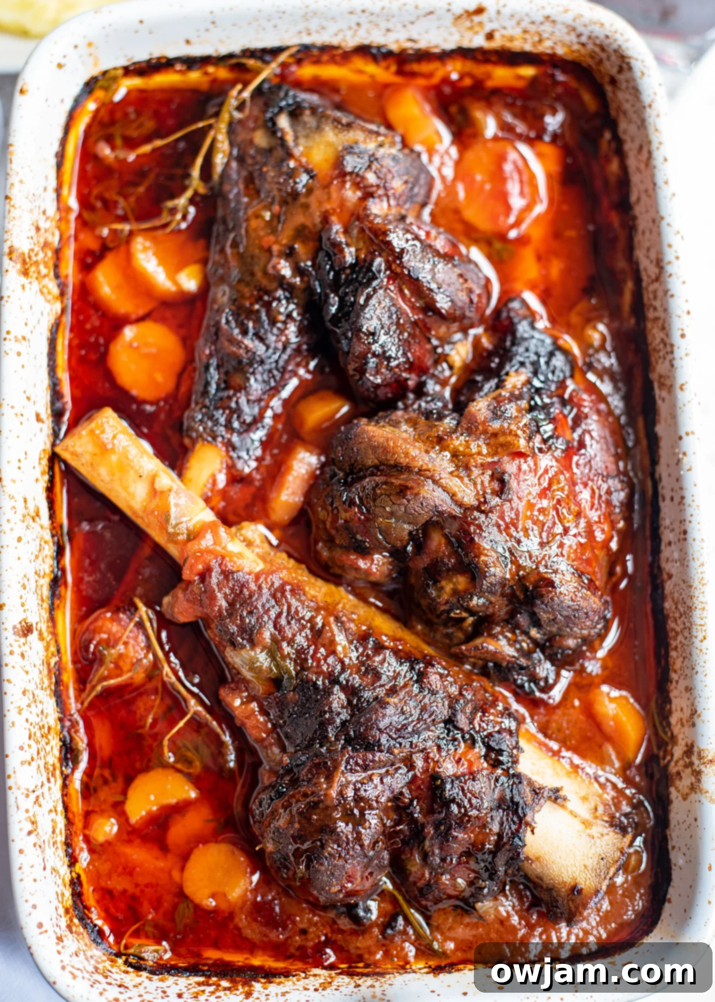 Braised lamb shanks simmering in a white baking dish with rich tomato sauce and vegetables.