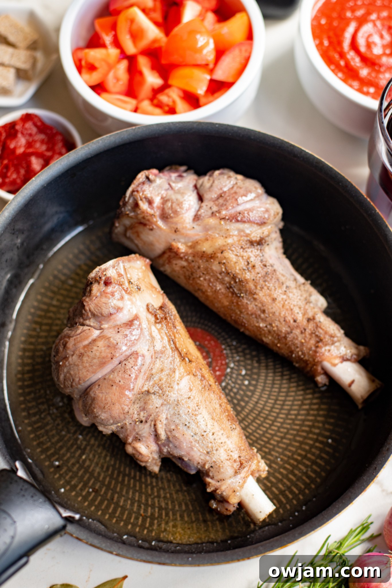Lamb shanks searing in a hot skillet with olive oil, browning beautifully.