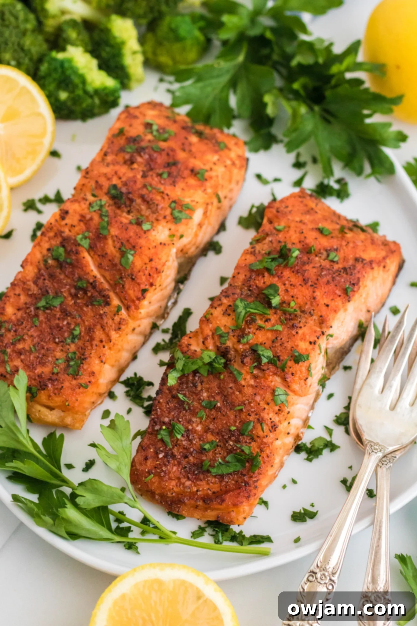Two seasoned and cooked salmon fillets garnished with chopped herbs are served on a white plate. Sliced lemons, parsley leaves, and broccoli florets surround the fillets. Two vintage-style forks are placed beside the salmon.