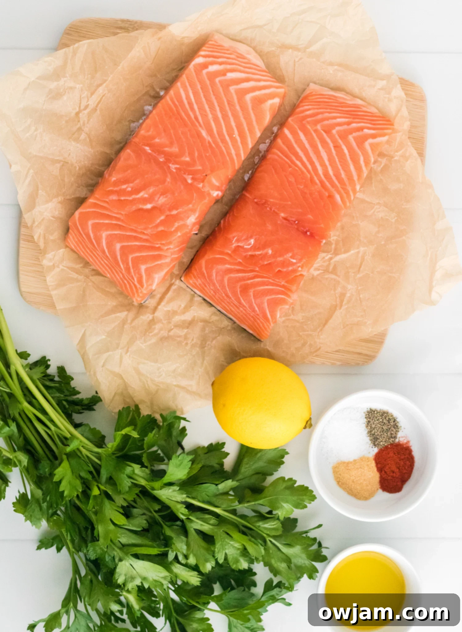 Two raw salmon fillets on parchment paper atop a wooden board. A bunch of fresh parsley, a lemon, and a small bowl containing spices, salt, and pepper are beside a bowl of olive oil on a white surface.
