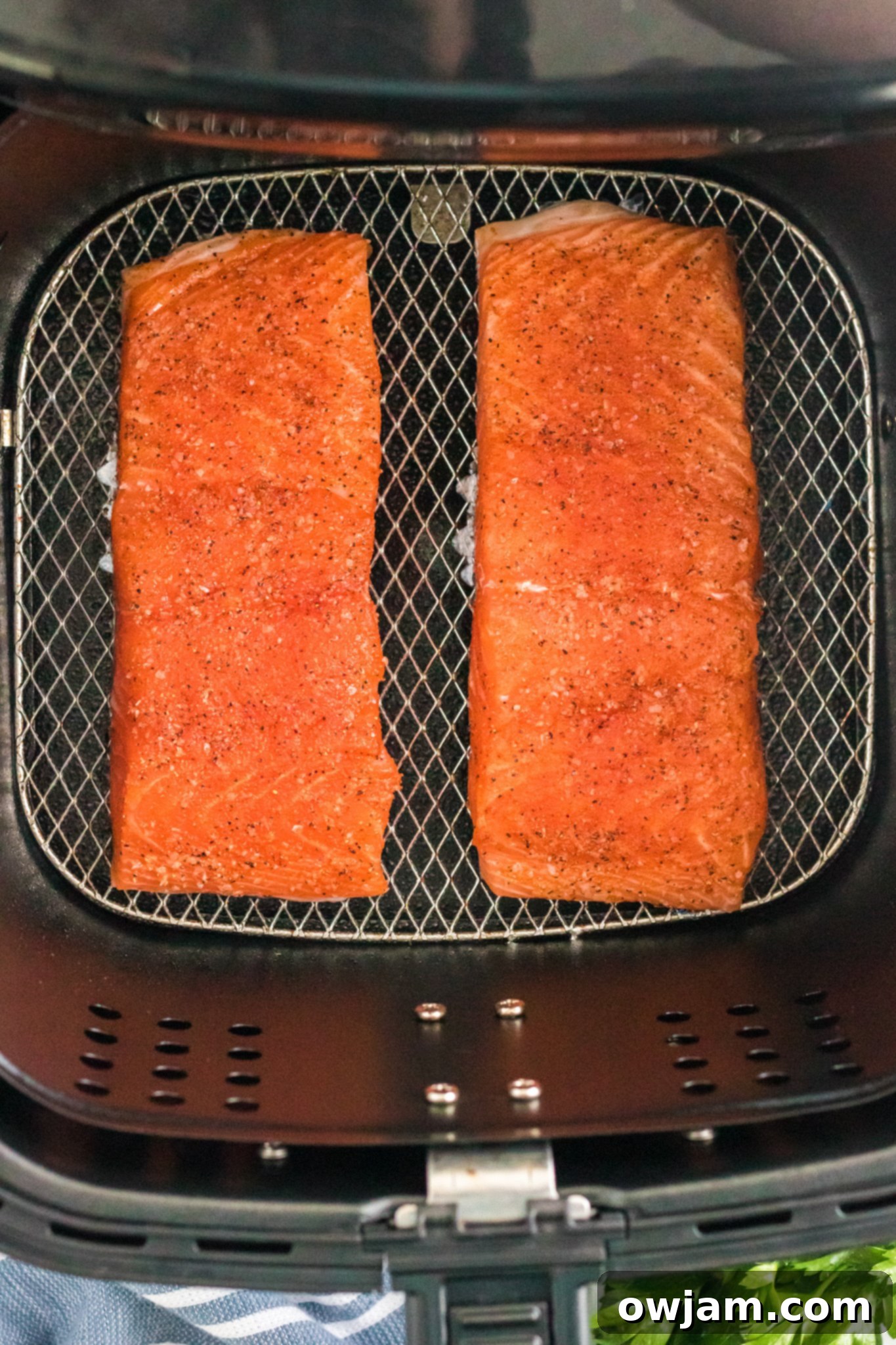 Two seasoned salmon fillets are placed side by side on the grill rack of an air fryer, ready for cooking. Fresh parsley and a blue-striped cloth can be seen below the open fryer basket.