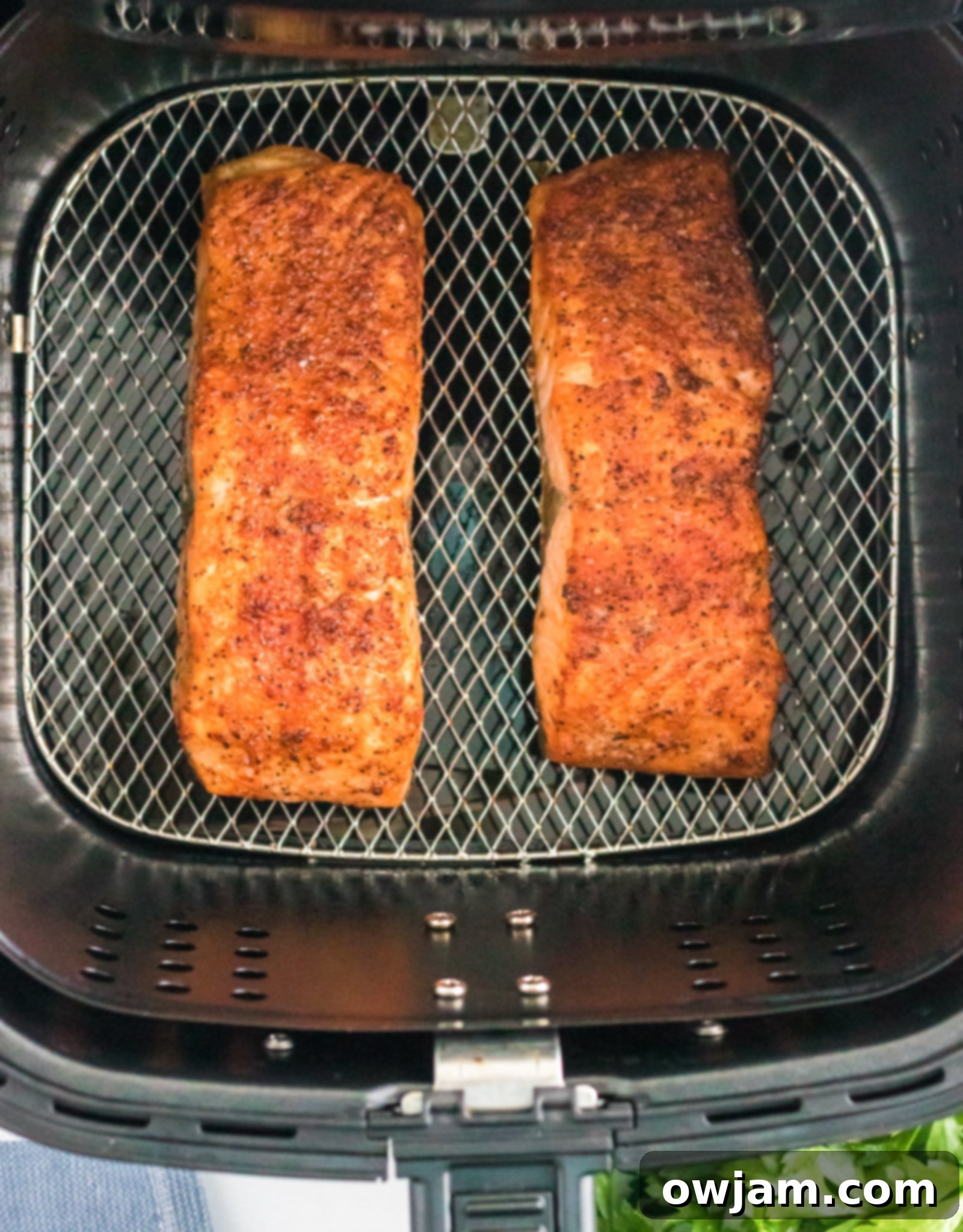 Two seasoned salmon fillets are cooking in an air fryer basket. The fillets are golden brown with a crispy texture. Fresh herbs are visible in the bottom right corner.