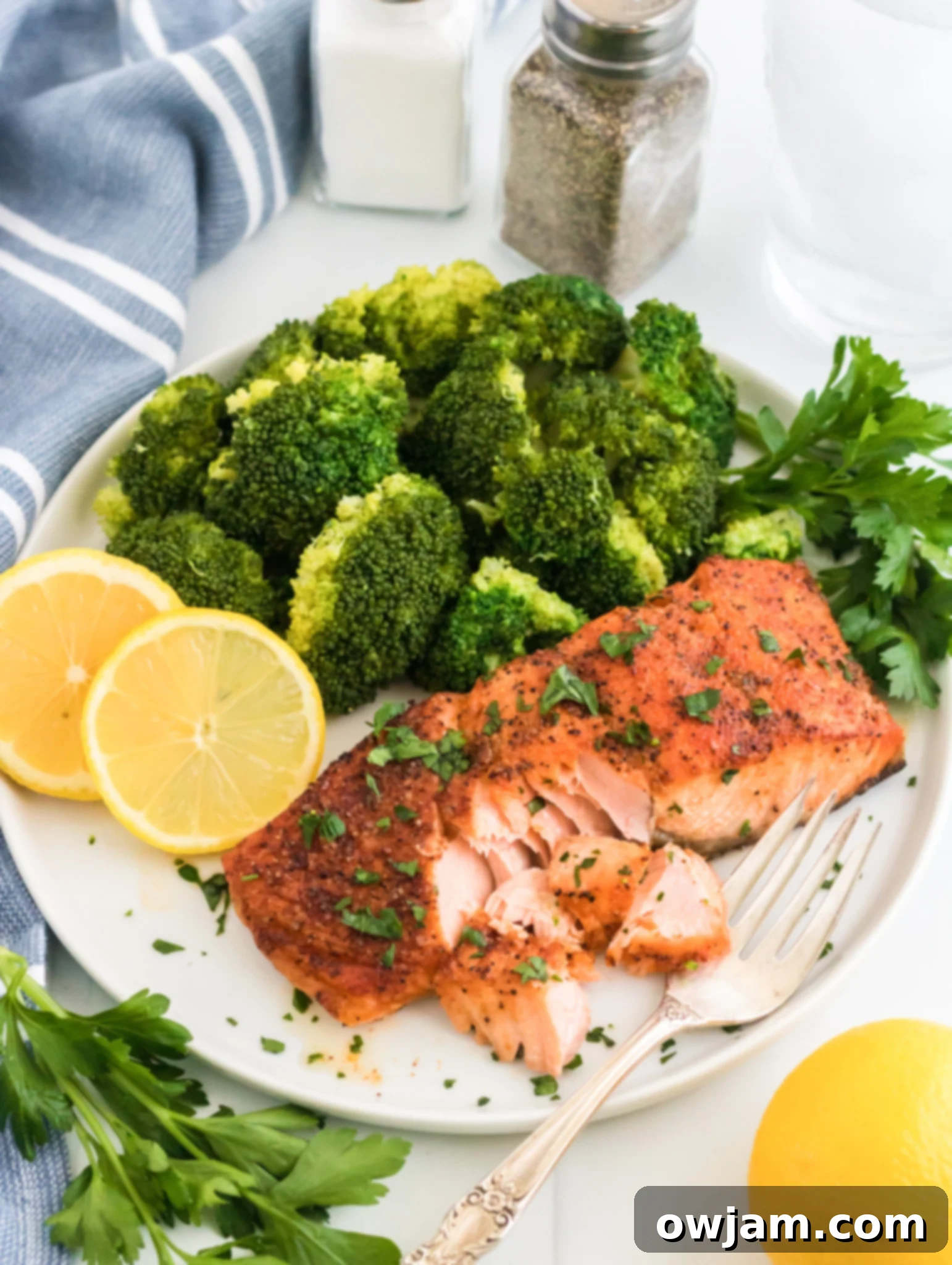 A plate with roasted salmon garnished with herbs, steamed broccoli, and lemon slices. A fork flakes the salmon. Salt and pepper shakers, a glass of water, and a striped cloth are in the background. Fresh parsley and a lemon are nearby.