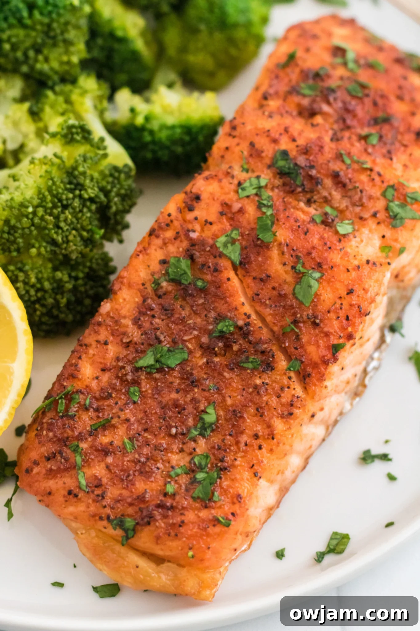 Grilled salmon fillet seasoned with herbs and pepper, garnished with chopped parsley, is served on a white plate alongside vibrant green broccoli florets and a lemon wedge.