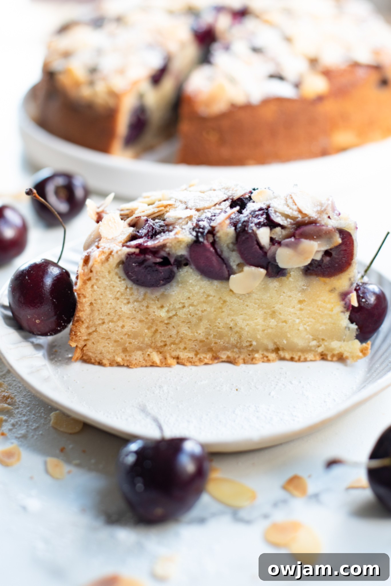 A large slice of Cherry Almond Cake with its perfect crumb, topped with juicy cherries and golden almonds, on a white plate.