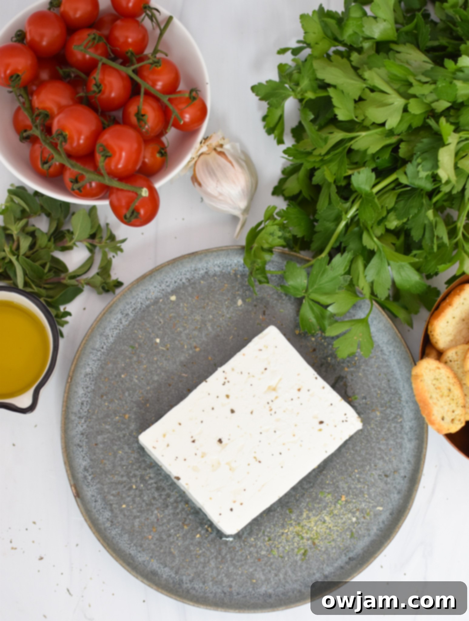 Simple Baked Feta Perfection 3 Ingredients for Easy Baked Feta Recipe laid out, including a block of feta cheese, fresh cherry tomatoes, olive oil, and various dried and fresh herbs.