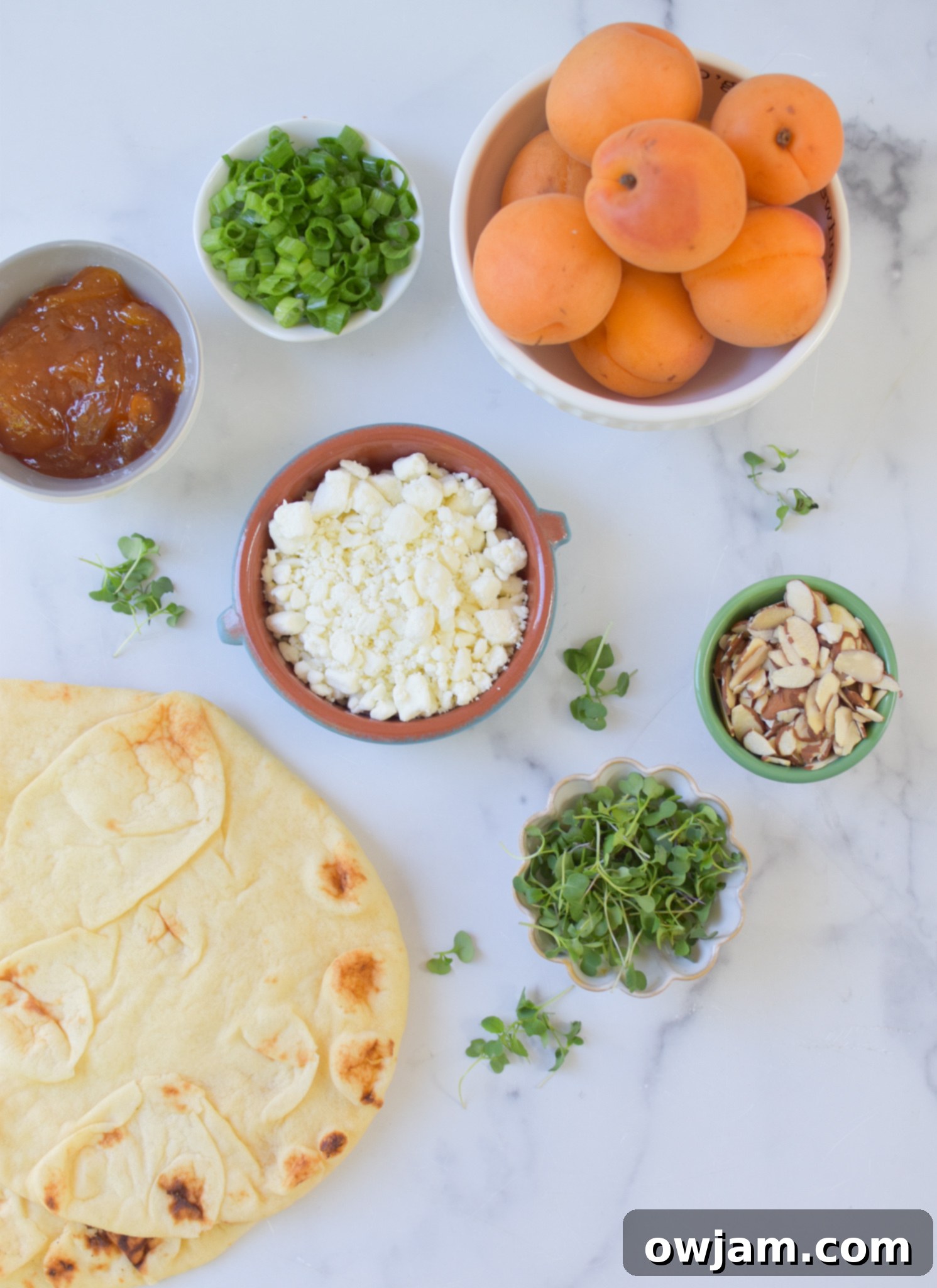 All ingredients for Goat Cheese Apricot Pizza laid out on a wooden board: naan bread, a jar of apricot jam, a container of goat cheese, sliced fresh apricots, sliced almonds, chopped green onions, and a bottle of honey.