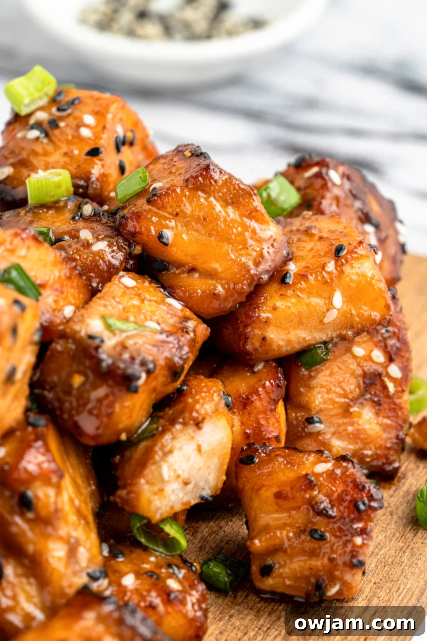 Crispy Teriyaki Air Fryer Salmon Bites 2 Close-up of glazed salmon cubes garnished with sesame seeds and green onions on a wooden board. A small bowl of sesame seeds is blurred in the background.