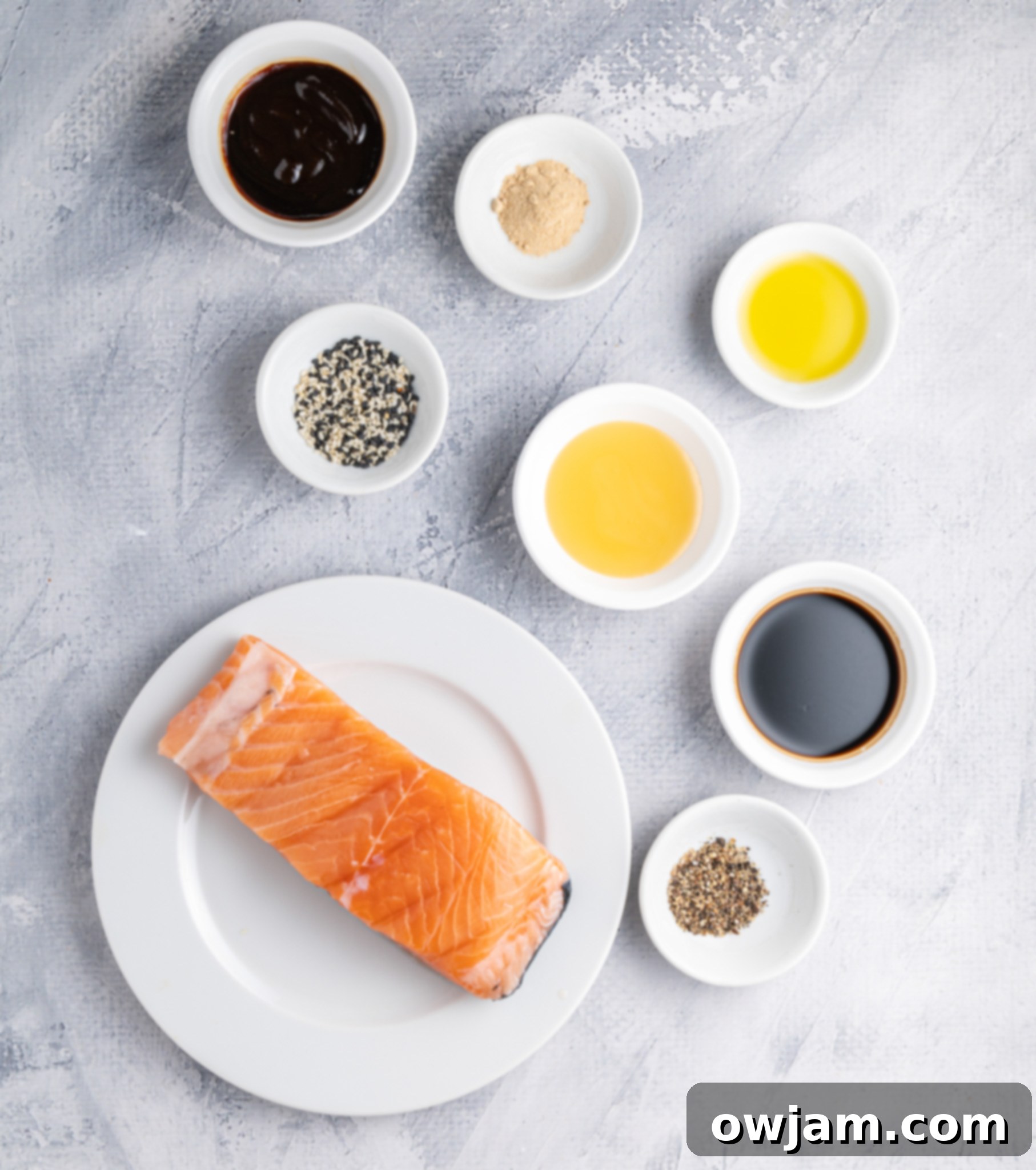 Crispy Teriyaki Air Fryer Salmon Bites 3 A salmon fillet on a white plate surrounded by small bowls containing various ingredients such as sauce, sesame seeds, oil, garlic powder, and pepper, arranged on a textured gray surface.