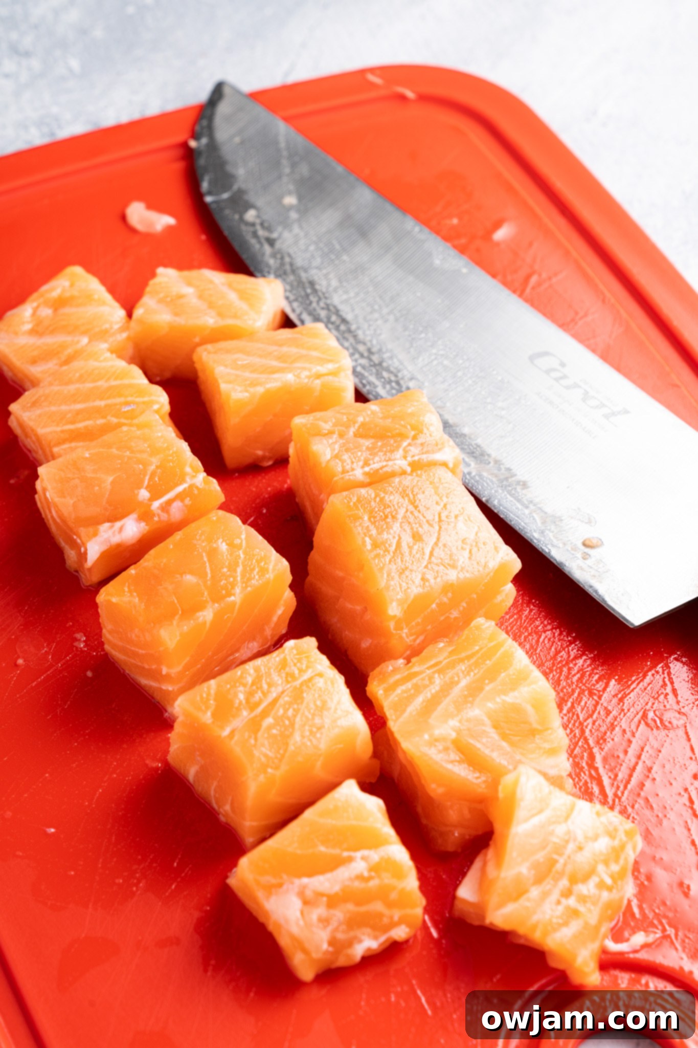 Crispy Teriyaki Air Fryer Salmon Bites 4 Cubes of fresh salmon are neatly arranged on a bright red cutting board. A large kitchen knife with a shiny blade is positioned next to the salmon pieces. The background has a textured, cool-toned surface.