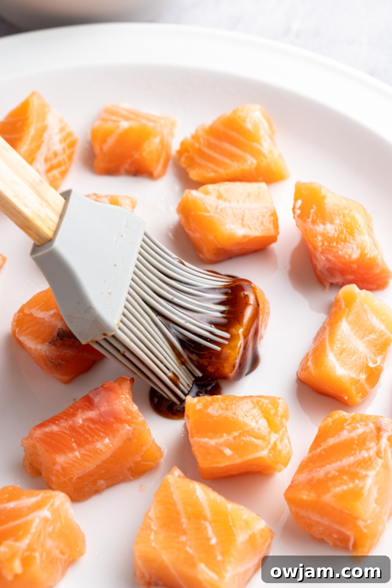 Crispy Teriyaki Air Fryer Salmon Bites 5 A silicone brush applies a dark marinade to raw salmon cubes on a white plate. The salmon pieces are evenly spaced and glistening, suggesting preparation for cooking. The background is softly blurred.
