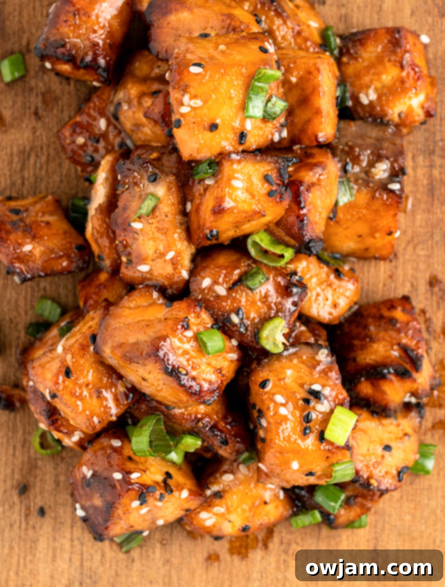 Crispy Teriyaki Air Fryer Salmon Bites 7 A pile of glazed, air fryer salmon bites topped with sesame seeds and sliced green onions on a wooden surface. The salmon has a caramelized appearance, indicating its well-cooked and flavorful.