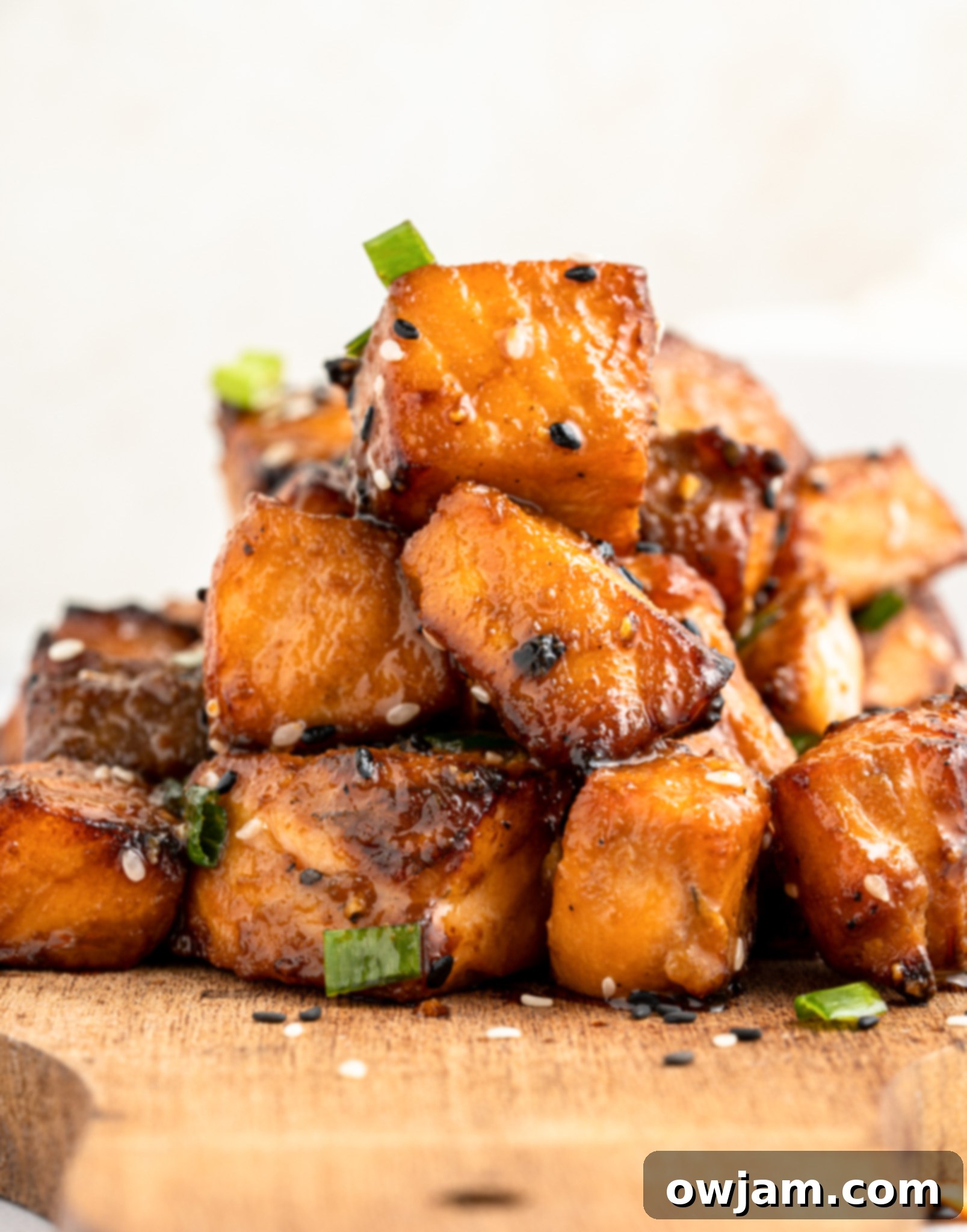 Crispy Teriyaki Air Fryer Salmon Bites 8 A stack of air fryer salmon bites garnished with sesame seeds and green onions on a wooden board. The pieces are golden-brown, highlighting their caramelized surface.