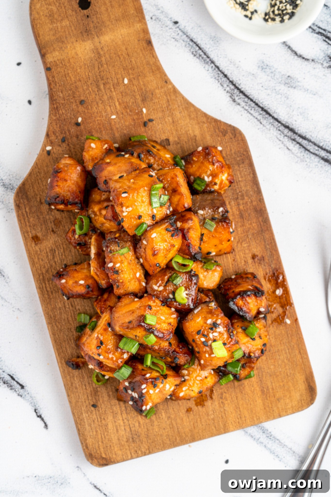 Crispy Teriyaki Air Fryer Salmon Bites 9 air fryer salmon bites glazed in a brown sauce, garnished with chopped green onions and sesame seeds, served on a wooden cutting board. A small dish with sesame seeds is in the background, set on a marble surface.