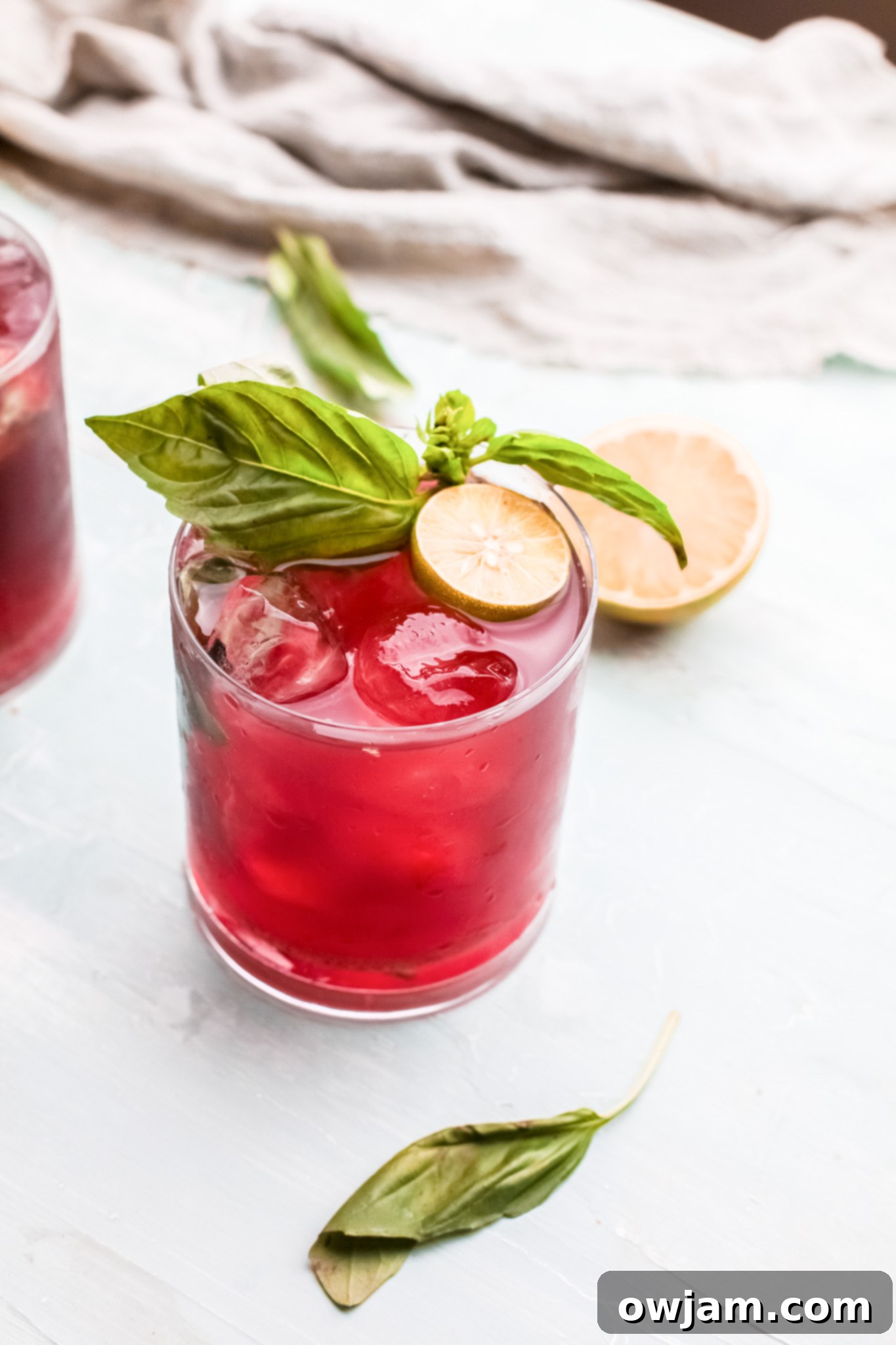 A close-up of a glass of Vodka Cherry Limeade, garnished with lime and basil, against a bright background.