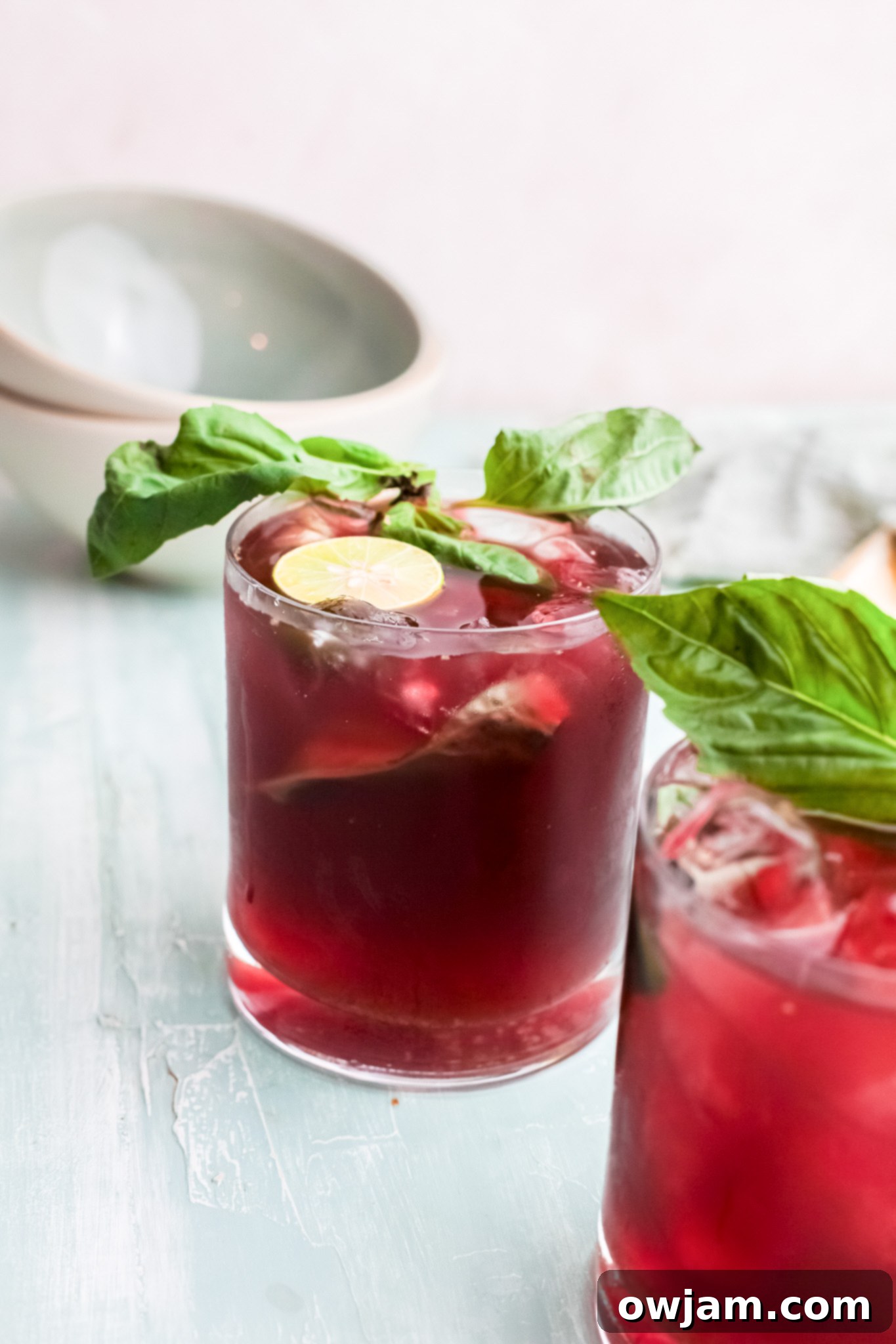 A vibrant Vodka Cherry Limeade in a tall glass, garnished with lime and basil, on a light background.