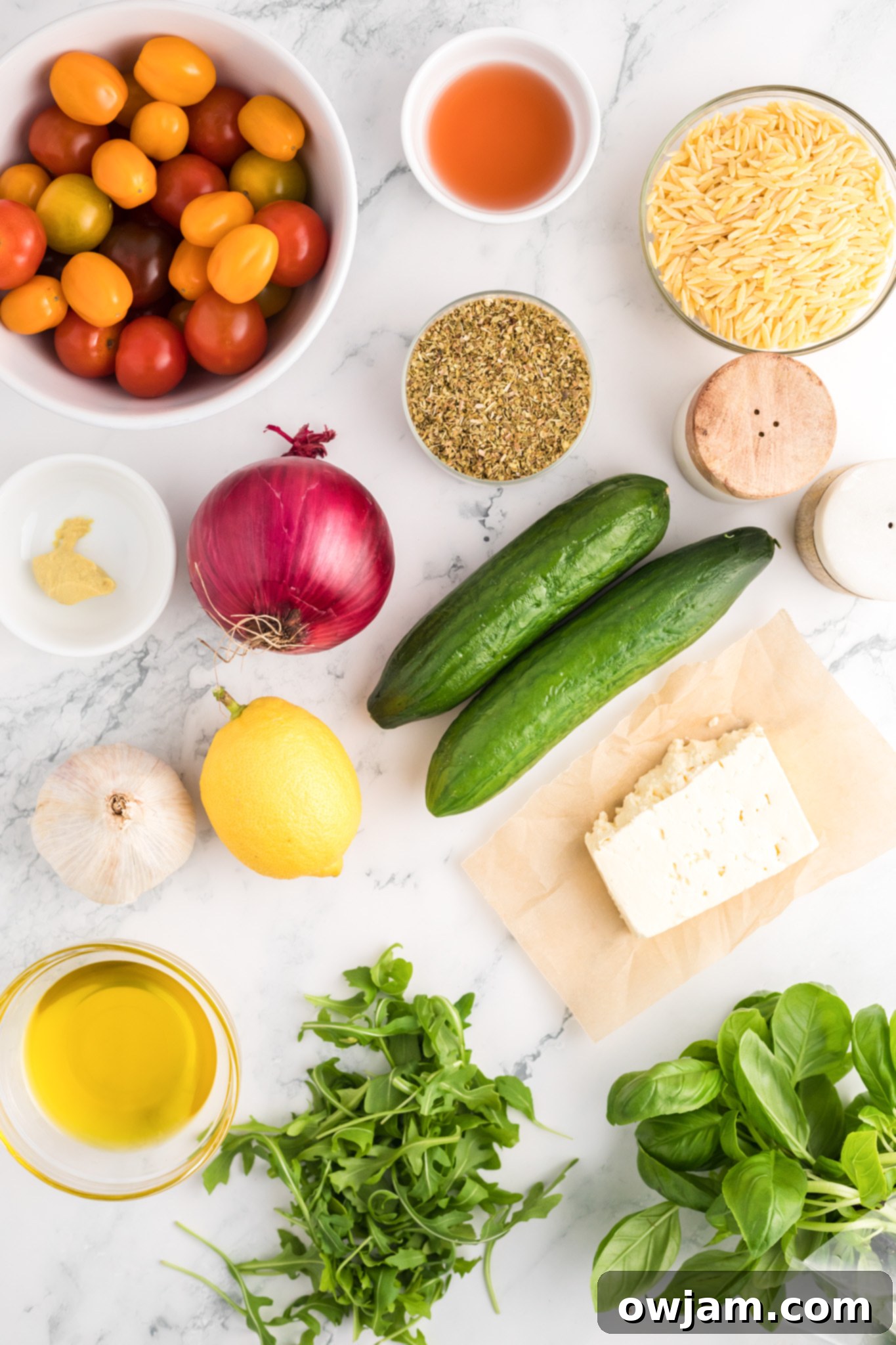 Various fresh ingredients for Arugula Orzo Salad laid out on a white surface, including orzo, arugula, tomatoes, cucumber, and feta.