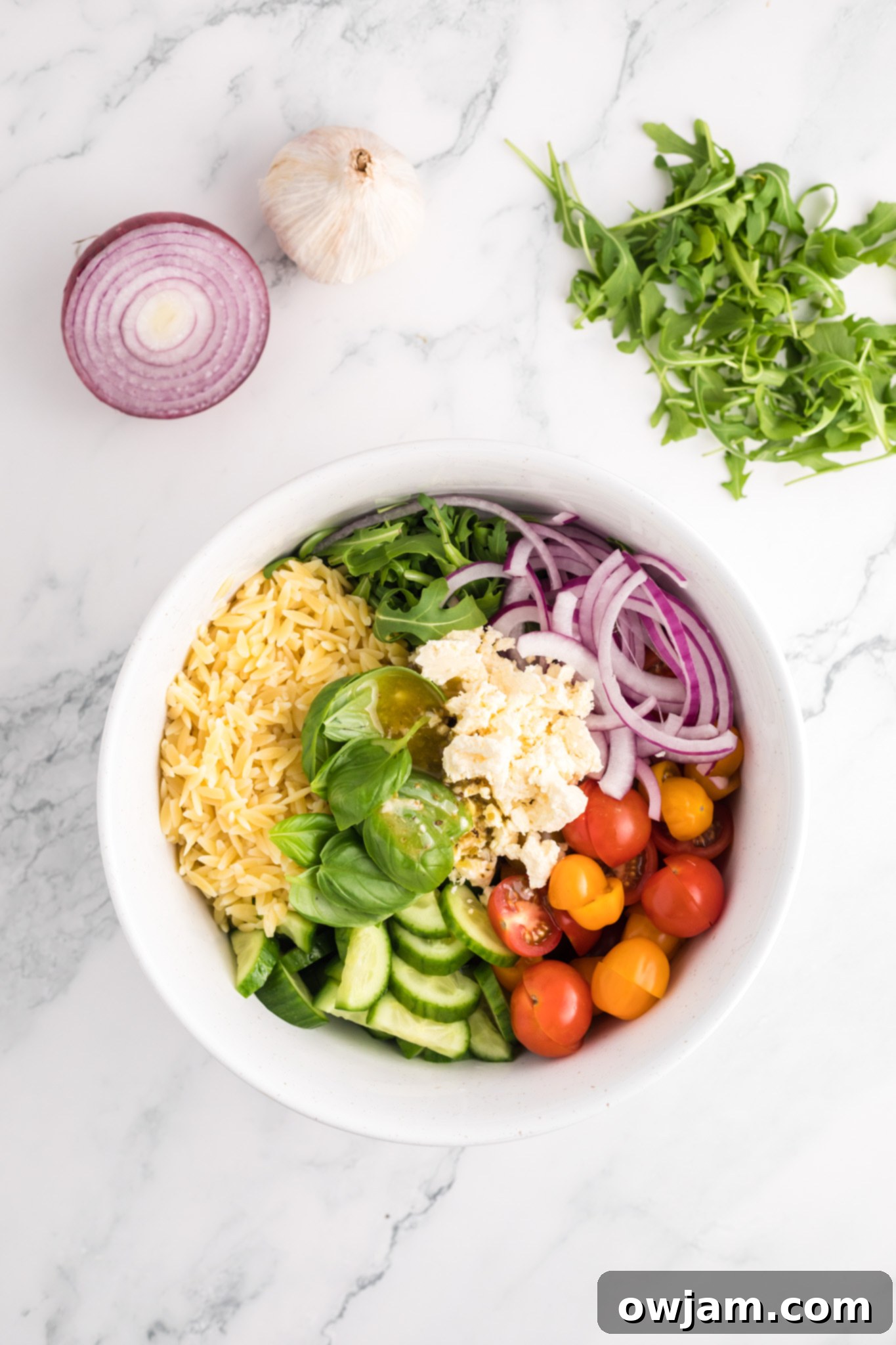 Fresh ingredients for Arugula Orzo Salad, including pasta, vegetables, and feta, combined in a large mixing bowl before dressing.