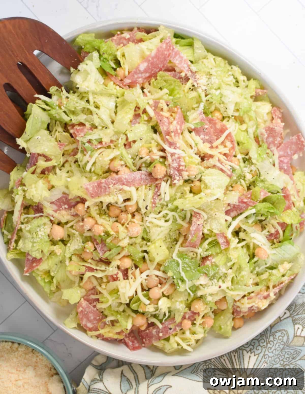 A large white bowl filled with Copycat La Scala Chopped Salad, featuring crisp lettuce, salami, cheese, and chickpeas, with wooden serving utensils resting inside.