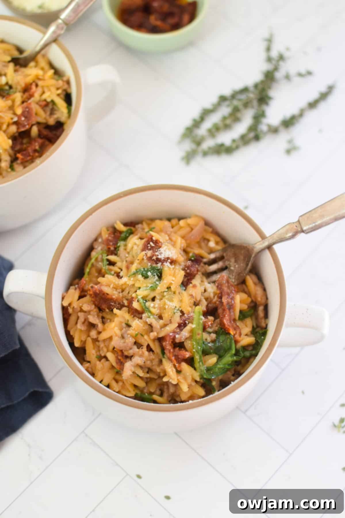 A small white bowl filled with creamy One Pot Sausage Orzo, garnished with fresh herbs, with a fork digging in.